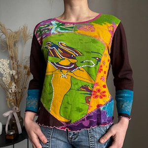 Könnte beinhalten: Ein langärmliges Patchwork-Oberteil mit einem lebendigen Design. Das Oberteil zeigt eine zentrale Grafik einer Figur in Gelb und Grün, akzentuiert mit weißen Perlendetails. Die Ärmel sind braun, mit blauen und türkisfarbenen Manschetten. Der Ausschnitt und der Saum sind rosa eingefasst.