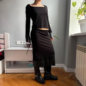 May include: A black long-sleeved top with a square neckline and matching black midi skirt. The top features black leather-like cuffs at the wrists, and the skirt has a wide black leather-like hem. The outfit is paired with black leather boots.
