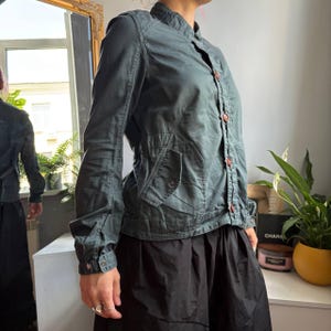 May include: A dark teal long-sleeved jacket featuring a mandarin collar, a front button closure with brown buttons, and two side pockets with flaps. The jacket has a structured fit with visible seam detailing along the torso and sleeves.