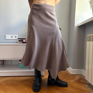 May include: A midi-length skirt in a muted mauve-gray tone, featuring a high-waisted fit and a flared, asymmetrical hemline with curved seam detailing. The skirt is paired with black leather boots that have silver-toned metal hardware accents.