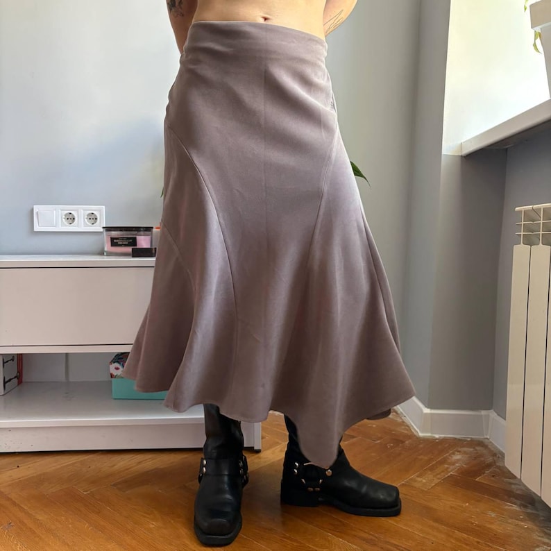 May include: A midi-length skirt in a muted mauve-gray tone, featuring a high-waisted fit and a flared, asymmetrical hemline with curved seam detailing. The skirt is paired with black leather boots that have silver-toned metal hardware accents.