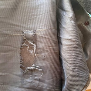 May include: Close-up of the torn lining inside a brown jacket. The lining is a light brown color with a striped pattern. The tear reveals the inner material of the jacket.