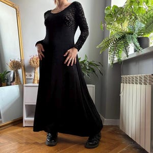 May include: A floor-length black dress featuring long sleeves and a scoop neckline. The bodice and sleeves are crafted from a textured, velvet-like fabric with a subtle tonal floral pattern, while the skirt is made of a smooth, solid black fabric.
