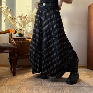 May include: A black and white striped maxi skirt with a high waist and a belt. The skirt has a flowing, flared design and a side slit. The person is wearing black boots.