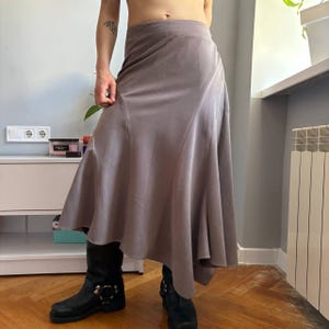 May include: A mauve-colored midi skirt featuring a high-waisted fit and a flared, asymmetrical hemline. The skirt is constructed with vertical panel seams that create a structured, flowing silhouette that falls below the knees.