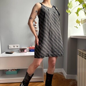 May include: A black and white patterned dress with thin straps and a bow detail. The dress has a diamond pattern and falls to mid-thigh. The person is wearing black boots. The dress is a summer or spring style.