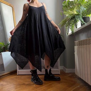 May include: A black sleeveless dress featuring a scoop neckline and thin shoulder straps. The garment has a loose, flowing silhouette with an asymmetrical handkerchief hemline and a detailed, intricate eyelet lace pattern throughout the fabric.