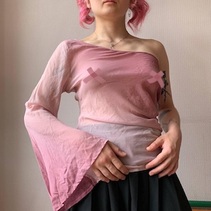 May include: A pink, one-shoulder top with a long, flared sleeve. The top has a gradient effect, transitioning from a darker pink to a lighter shade. The fabric appears lightweight and slightly sheer, with a draped design. The bottom is a black skirt.
