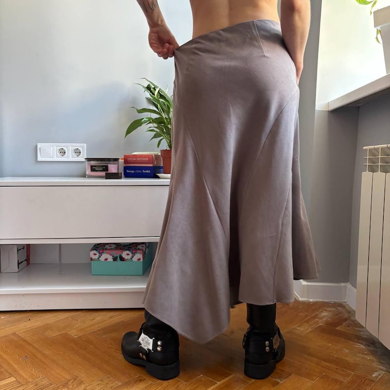May include: A midi-length skirt in a muted taupe color featuring a smooth, matte finish and a flared, asymmetrical hemline. The skirt has a high-waisted fit with visible vertical seam detailing that creates a structured, flowing silhouette.