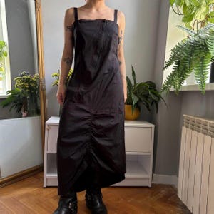 May include: A black sleeveless midi dress featuring a square neckline, thick shoulder straps, and a central vertical ruched detail extending from the waist to the hem, creating a gathered texture throughout the front of the garment.