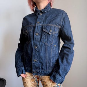 Puede incluir: Chaqueta vaquera azul oscuro con cierre de botones, dos bolsillos en el pecho y dos bolsillos laterales. La chaqueta tiene un cuello clásico y está hecha de un material de mezclilla resistente. La chaqueta tiene un corte entallado.