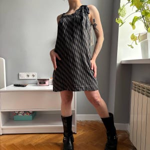 May include: A black and white patterned dress with a unique shoulder tie detail. The dress is knee-length and has a geometric design. The model is wearing black boots. The dress is a stylish choice for a casual or semi-formal occasion.