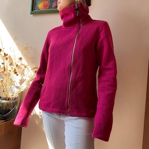 Puede incluir: Chaqueta de forro polar magenta con cuello alto y cremallera plateada completa. La chaqueta tiene mangas largas y un aspecto texturizado. El modelo lleva pantalones blancos. Una prenda de abrigo cálida y cómoda.