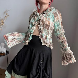 Könnte beinhalten: Eine transparente, offen geschnittene Bluse mit Rüschenkragen und Ärmeln. Die Bluse hat ein Blumenmuster in Blau-, Braun- und Rosatönen. Das Model trägt einen schwarzen Rock.