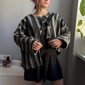May include: A gray and black striped jacket with a unique toggle closure. The jacket has wide sleeves and a textured fabric. The model is wearing a black skirt. The jacket is a stylish outerwear option.