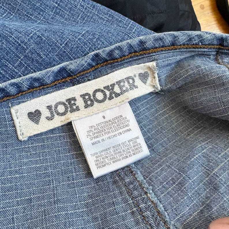 Może przedstawiać: Zbliżenie niebieskiego dżinsowego ubrania z metką z napisem "JOE BOXER" w szarym tekście. Etykieta zawiera r&oacute;wnież numer "9" oraz szczeg&oacute;ły dotyczące składu materiału. Widoczna jest faktura ubrania, ze szwami i brązowym szwem.