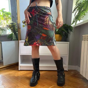 Op de afbeelding: Een rok tot op de knie met een donker, veelkleurig patchworkpatroon met groene, oranje en paarse geometrische vormen. De rok heeft een taille met blauwe trekkoorden en een klein wit geborduurd logo bij de onderste zoom.