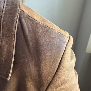 May include: Close-up of a brown leather jacket shoulder. The leather has a distressed appearance with visible grain and slight imperfections. The collar and shoulder seam are visible, showcasing the jacket's construction.