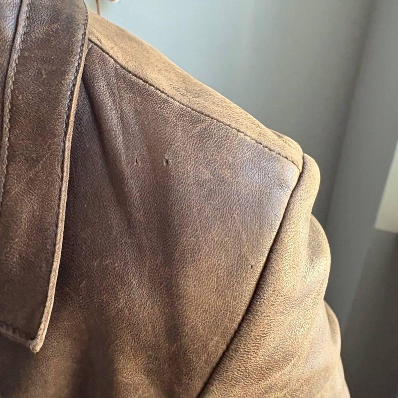 May include: Close-up of a brown leather jacket shoulder. The leather has a distressed appearance with visible grain and slight imperfections. The collar and shoulder seam are visible, showcasing the jacket's construction.