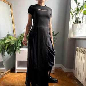 May include: A black floor-length dress with short sleeves, a horizontal cutout detail across the chest, and a high-waisted skirt featuring a long vertical slit that reveals a draped, layered fabric panel underneath the main skirt structure.