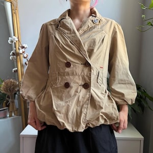May include: A tan double-breasted jacket featuring a wide collar, three-quarter length puff sleeves with gathered cuffs, and four dark brown buttons on the front. The jacket has a cinched waistline and a slightly flared, gathered hemline.