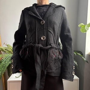 May include: A dark charcoal grey jacket featuring a hood, a front button closure with two large circular buttons, and a matching fabric tie belt at the waist. The jacket includes long sleeves with two buttons at each cuff and decorative red stitching.