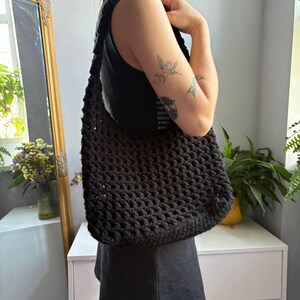 May include: A black crochet shoulder bag featuring a thick, textured yarn construction with an open-knit pattern. The bag has a wide, integrated strap and a rectangular body shape, designed to be worn over the shoulder with a soft, flexible structure.