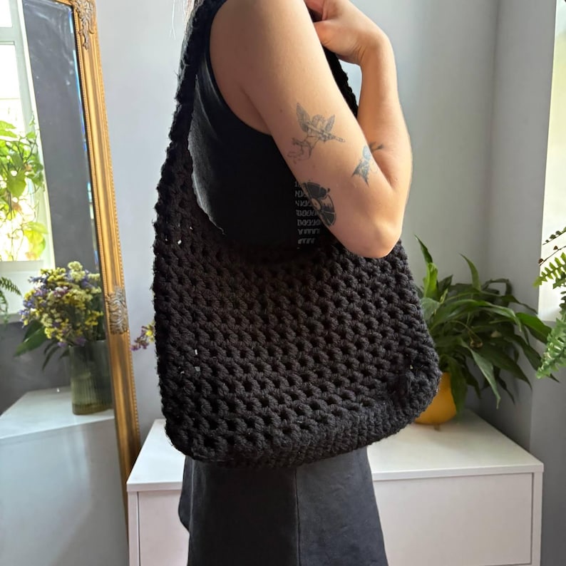 May include: A black crochet shoulder bag featuring a thick, textured yarn construction with an open-knit pattern. The bag has a wide, integrated strap and a rectangular body shape, designed to be worn over the shoulder with a soft, flexible structure.