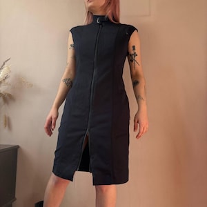 Puede incluir: Vestido negro sin mangas con cuello alto y cremallera completa en la parte delantera. El vestido tiene un dobladillo a la altura de la rodilla y un corte entallado. El vestido está hecho de una tela oscura y lisa.
