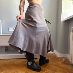 May include: A long, taupe-colored skirt with a smooth, matte texture and a flared, panelled design that creates a flowing silhouette. The skirt features vertical seams that extend from the waist down to the hem, providing a structured shape.
