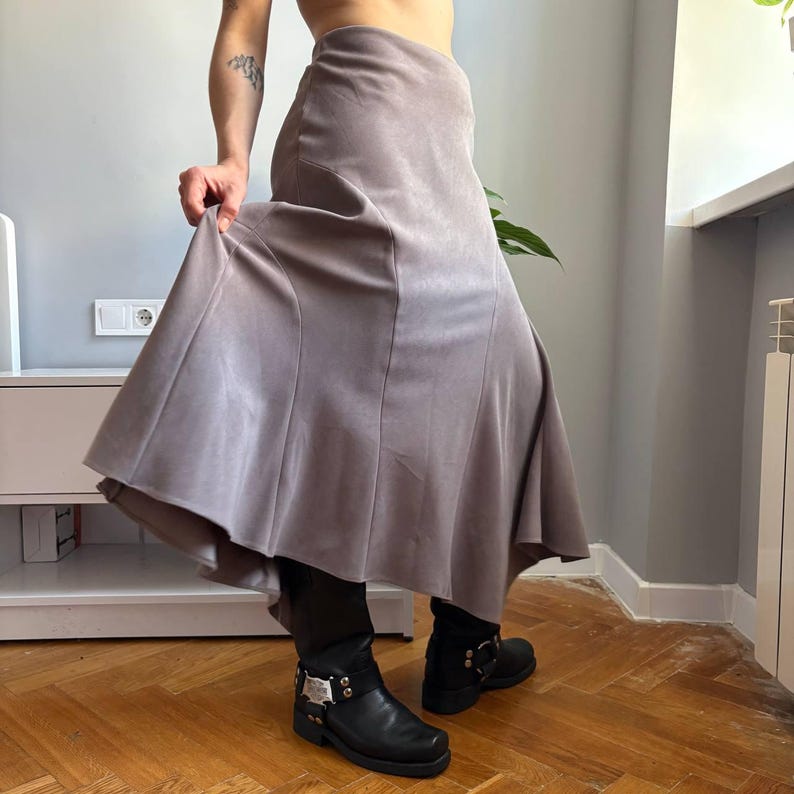 May include: A long, taupe-colored skirt with a smooth, matte texture and a flared, panelled design that creates a flowing silhouette. The skirt features vertical seams that extend from the waist down to the hem, providing a structured shape.