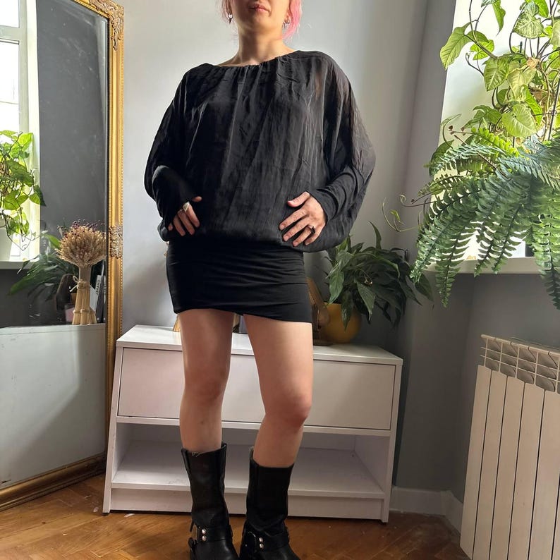 May include: A black long-sleeved top with a loose, draped bodice and a gathered neckline, paired with a fitted black mini skirt and black mid-calf boots featuring silver hardware accents. The ensemble consists of a sheer, lightweight blouse.