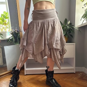 May include: A taupe-colored skirt featuring a wide, flat waistband and a multi-layered, handkerchief-style hemline. The fabric has a subtle, crinkled texture, and the skirt falls in uneven, draped panels that create a voluminous, layered appearance.