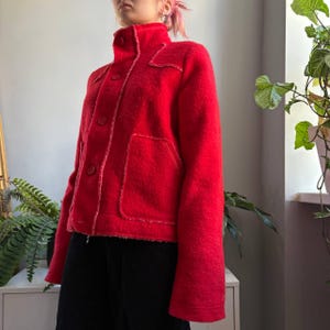 May include: A vibrant red jacket featuring a high collar, a front button closure, and two large patch pockets. The garment is constructed with visible, raw-edge seams along the placket, pockets, and shoulder panels, providing a textured finish.