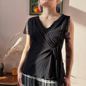 May include: Black sleeveless top with a V-neck and lace detailing on the right shoulder. The top has a draped design and is paired with a black and white plaid skirt. The top is made of a soft, flowing fabric.