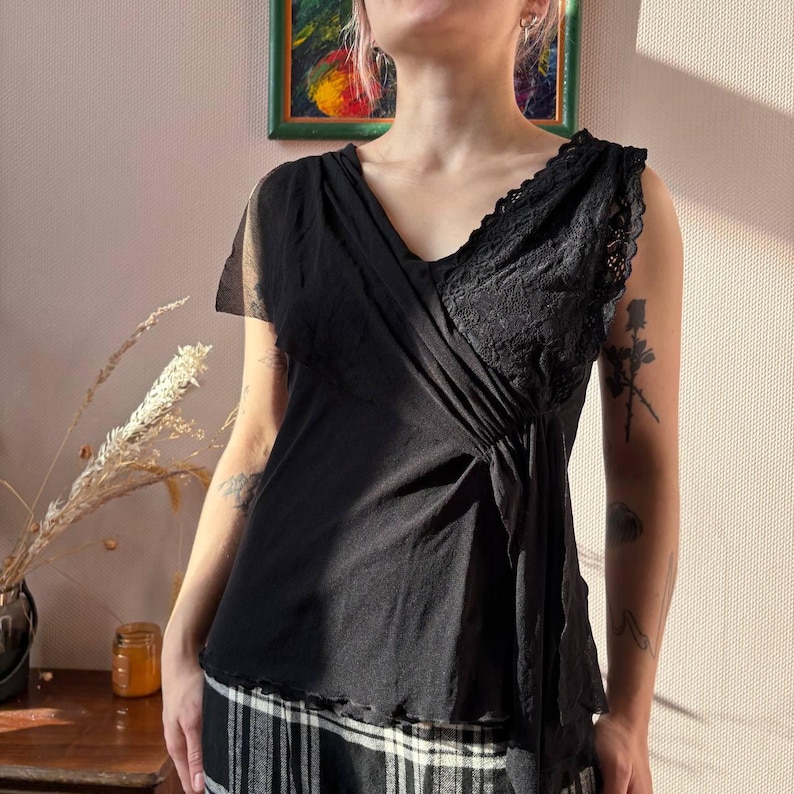 May include: Black sleeveless top with a V-neck and lace detailing on the right shoulder. The top has a draped design and is paired with a black and white plaid skirt. The top is made of a soft, flowing fabric.