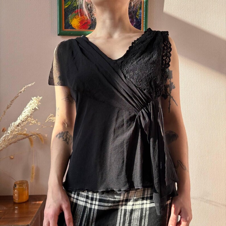 May include: Black V-neck top with lace detailing on the right shoulder. The top has a draped design and short sleeves. The model is wearing a black and white plaid skirt. The background includes a painting and a jar.