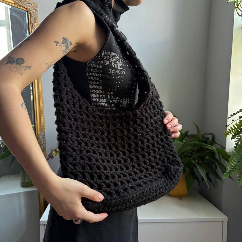 May include: A black crochet shoulder bag featuring a thick, textured yarn construction with an open-knit pattern. The bag has a wide, integrated strap and a rectangular body shape, crafted from a solid black fiber with a consistent, repeating stitch.