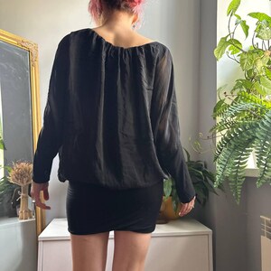 May include: A black long-sleeved blouse with a gathered neckline and a loose, draped fit, paired with a fitted black mini skirt. The blouse features sheer, lightweight fabric on the sleeves and a solid, opaque bodice with a cinched hemline.