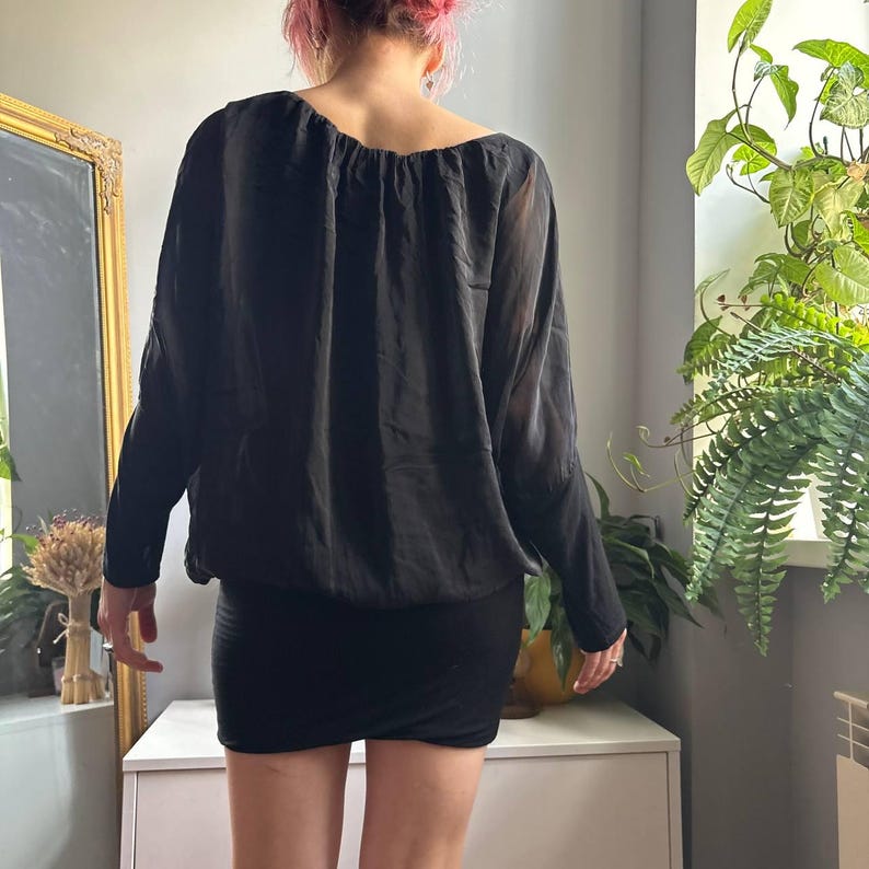 May include: A black long-sleeved blouse with a gathered neckline and a loose, draped fit, paired with a fitted black mini skirt. The blouse features sheer, lightweight fabric on the sleeves and a solid, opaque bodice with a cinched hemline.