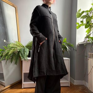 May include: A long-sleeved black coat featuring a high collar, a full-length silver-toned front zipper, and two side pockets. The garment has a textured fabric finish and a flared, asymmetrical hemline that falls below the hips.