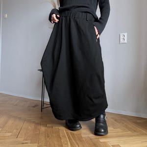 May include: A long, black skirt with a wide waistband and pockets. The skirt has a flowing, asymmetrical design and is paired with black boots. The outfit is simple and modern, suitable for various occasions.