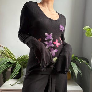 Puede incluir: Una camiseta negra de manga larga con cuello redondo y puños con volantes. La parte delantera de la prenda presenta un patrón floral decorativo de fieltro con aguja en tonos morado, rosa y verde, creando un diseño texturizado sobre la tela.