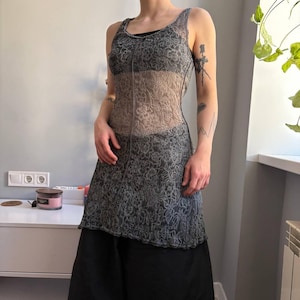 May include: A sleeveless dress featuring a gray floral lace overlay. The dress has a sheer panel down the center and a black skirt underneath. The model is wearing a black top under the dress. The dress is a unique piece of clothing.