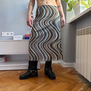 Puede incluir: Falda larga con un patrón ondulado en tonos marrón, blanco y negro. La falda se combina con botas de cuero negras. El diseño de la falda crea un ambiente retro y psicodélico.