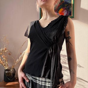 May include: Black sleeveless top with a V-neckline and lace detailing. The top features a draped design across the front and a layered, asymmetrical hem. The fabric appears to be a combination of soft, flowing material and delicate lace.
