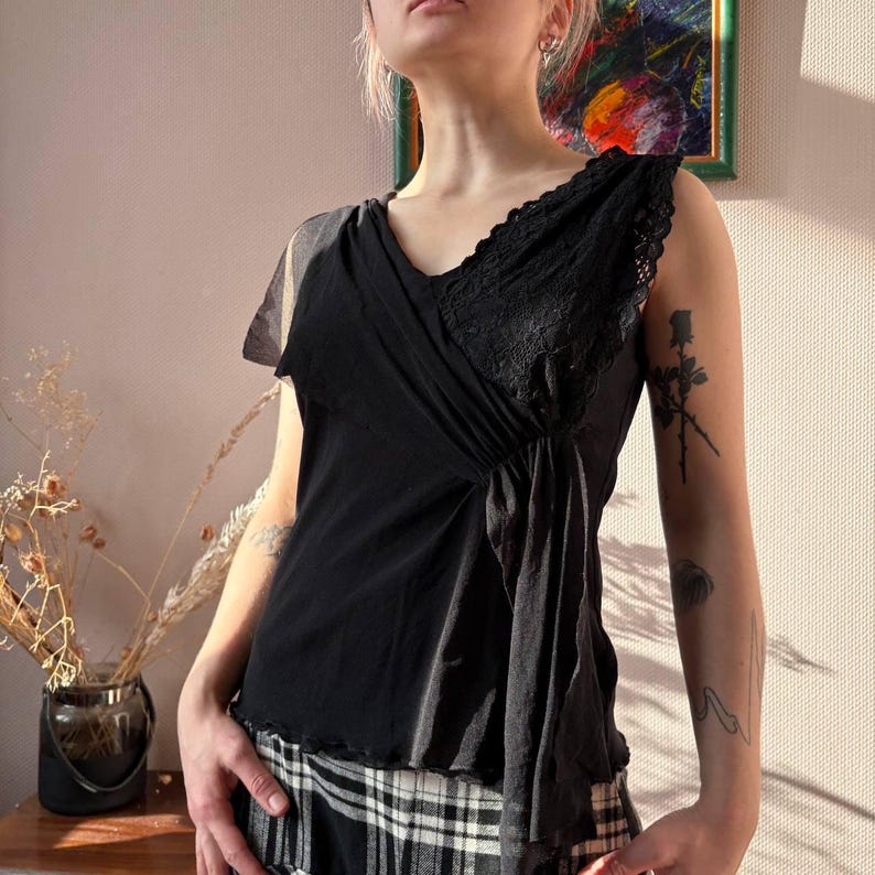 May include: Black sleeveless top with a V-neckline and lace detailing. The top features a draped design across the front and a layered, asymmetrical hem. The fabric appears to be a combination of soft, flowing material and delicate lace.
