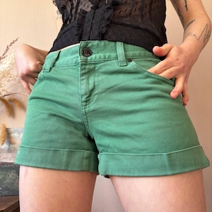Puede incluir: Shorts vaqueros verdes con dobladillo vuelto. Los shorts tienen cierre de botón y bolsillos. La tela parece ser de color verde medio liso. Los shorts son de estilo casual.