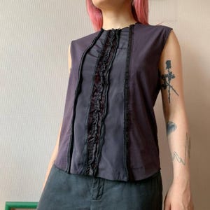 May include: Sleeveless top in shades of purple and grey. The top features a central panel with ruffled detailing and vertical accents. The design includes a mix of textures and tones, creating a layered effect. The garment has a simple, modern silhouette.