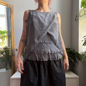 May include: A sleeveless grey top featuring a layered, asymmetrical design with raw-edge fabric panels that overlap across the front. The garment has a round neckline and a textured, slightly wrinkled appearance, paired with a solid black skirt.